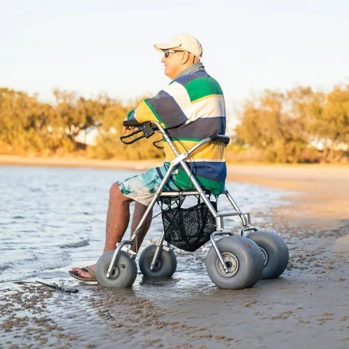 An elderly man sits on a rollator walker with large wheels designed for rough terrain, positioned at the edge of a sandy beach. He is dressed in a colourful striped shirt, blue and white tie-dye shorts, and a light-coloured cap, and wears sunglasses. The beach and calm water create a tranquil backdrop with trees visible in the distance.