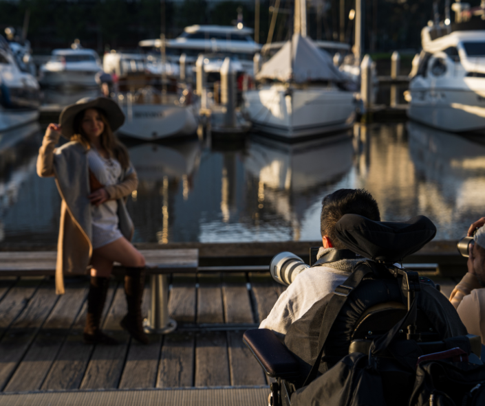 A photographer in a wheelchair, equipped with a large camera and telephoto lens, captures a photo of a woman posing on a wooden dock. The dock is adjacent to a marina filled with various boats and yachts, with calm water reflecting the vessels and creating a serene atmosphere. The woman, wearing a wide-brimmed hat, a light dress, and knee-high boots, stands confidently against the picturesque backdrop.