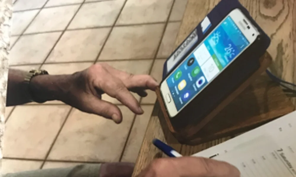 "A close-up image of an elderly person's hand interacting with a smartphone that is placed on a wooden stand. The screen of the phone displays several app icons and the weather. The person is using their index finger to touch the screen, and there is a pen and paper nearby on the table. The background includes a tiled floor, indicating an indoor setting.