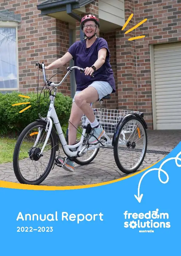 Cover page of the Annual Report 2022–2023 for Freedom Solutions Australia. The top half features a smiling woman wearing a helmet, riding a white tricycle in front of a brick house. Yellow lines around her indicate joy and excitement. The bottom half of the cover is blue with the Freedom Solutions Australia logo on the right, and the text "Annual Report 2022–2023" on the left. A white arrow with a curly line points from the tricycle to the logo.