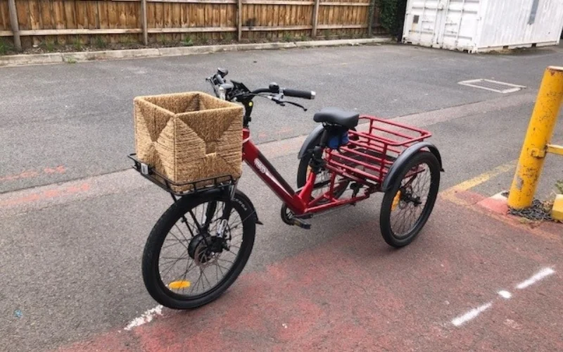 A red adaptive tricycle is parked on a paved surface in an industrial or suburban area. It features a woven basket on the front and a larger metal basket on the rear, providing ample storage space. The tricycle is designed for stability and ease of use, with three wheels and a comfortable seat. The background includes a wooden fence and a white storage container, indicating an outdoor setting with some industrial elements."