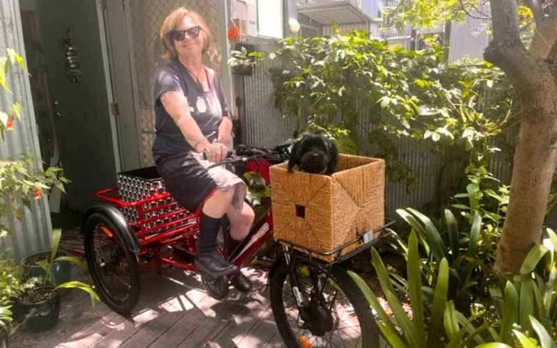 A woman sits on a red adaptive tricycle in a garden setting. She is wearing sunglasses and a dark outfit, appearing relaxed and content. The tricycle has a front woven basket with a small black dog inside, and a larger metal basket at the back. The background includes lush green plants and a tree, suggesting a peaceful, outdoor environment. The scene conveys a sense of independence and enjoyment of nature.
