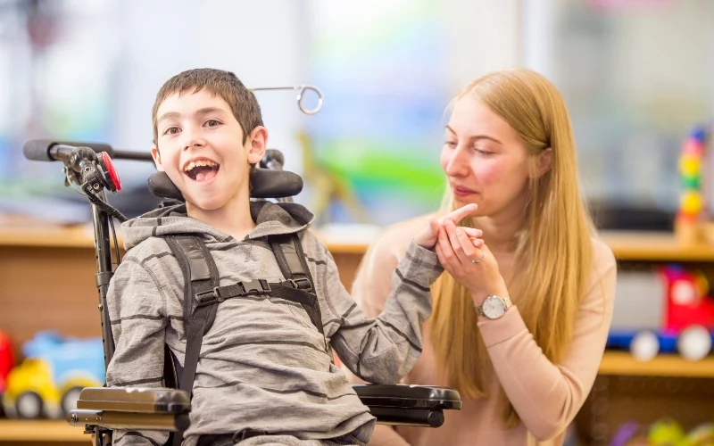 A young boy with a bright smile sits in a specialised wheelchair, holding hands with a woman kneeling beside him. The woman, possibly a caregiver or therapist, looks at him with a warm and supportive expression. The background is softly blurred, featuring colourful toys and a well-lit room, indicating a positive and engaging environment. The image radiates joy, connection, and the supportive care provided to children with disabilities