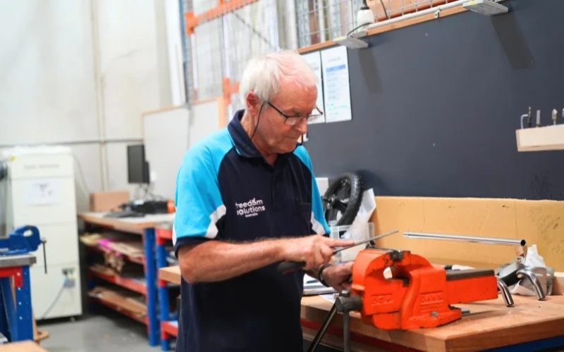 A senior man works diligently at a workbench in a workshop. He wears a Freedom Solutions Australia polo shirt and safety glasses, focusing intently on a piece of equipment held in a bright orange vice. The workshop background is organised with tools and materials neatly arranged, reflecting a professional and efficient workspace. The image conveys dedication and skilled craftsmanship, highlighting the technical expertise of the team at Freedom Solutions Australia.