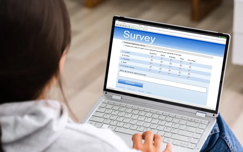 A person seated at a table or desk uses a laptop to complete an online survey. The screen displays a survey form with multiple-choice questions and rating scales. The individual is viewed from behind, focusing on the task at hand, indicating active participation in providing feedback or data. The setting appears to be a home or casual environment, suggesting convenience and accessibility for completing the survey.