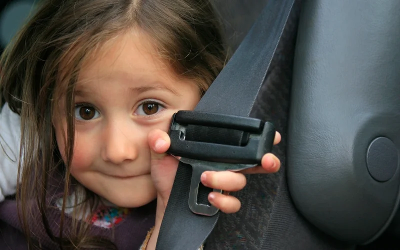 "A close-up image of a young girl with long brown hair and big brown eyes, peeking out from behind a seatbelt in a car. She holds the seatbelt latch with her hand, displaying a curious and playful expression. The background shows the car seat and interior, creating a sense of safety and protection."