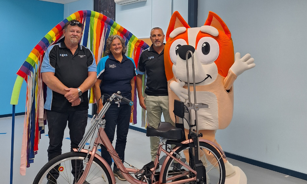 Three individuals stand in a brightly coloured room, smiling at the camera. Two of them wear shirts with the logos 'C4K' and 'Freedom Solutions Australia.' They are positioned beside a custom-built tricycle. A person in a large, cheerful cartoon character costume is also present, adding a playful element to the scene. The background features a rainbow arch made of fabric strips, enhancing the vibrant atmosphere.