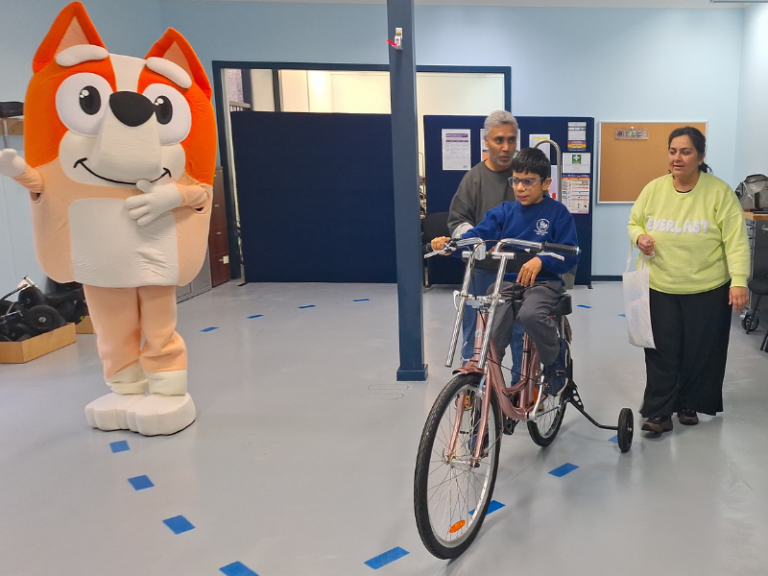 A child rides a custom-built tricycle indoors, supported by two adults walking alongside. A person in a cheerful cartoon character costume stands nearby, adding a playful and encouraging presence to the scene. The background shows a well-organised and spacious room, indicating a safe and controlled environment for practising and enhancing mobility skills. The overall atmosphere is supportive and positive, focused on promoting independence and confidence for the child.