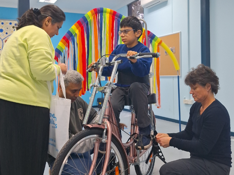 A child sits on a custom-built tricycle while three adults assist with adjustments and fittings. The scene takes place in a brightly decorated room, featuring a colourful rainbow arch made of fabric strips in the background. The child appears focused and engaged, holding the handlebars as the adults ensure the tricycle is properly configured for safety and comfort. The setting conveys a supportive and collaborative environment dedicated to enhancing mobility and independence for children with disabilities.