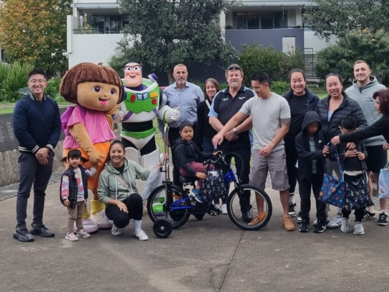 "A group of people, including children and adults, gather outdoors for a joyful event. Among them are individuals in colourful character costumes, including Dora the Explorer and Buzz Lightyear, adding a fun and engaging element to the scene. The group centres around a child sitting on an adaptive tricycle, with various members smiling and interacting. The background features greenery and modern buildings, creating a pleasant and lively atmosphere.