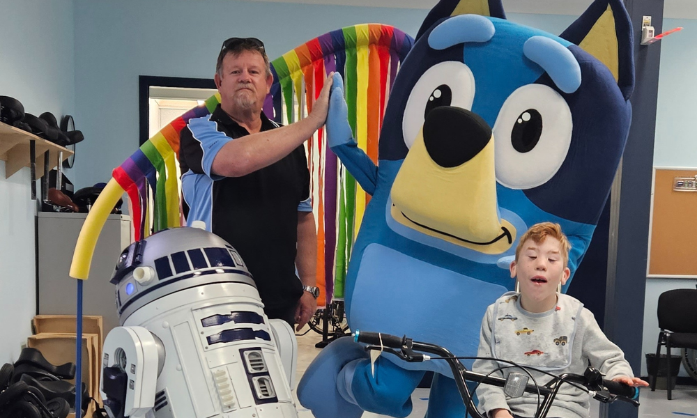 A young boy on a tricycle poses with a man giving a high-five to a person in a large Bluey character costume, next to an R2-D2 replica robot, under a colourful ribbon arch. The scene is playful and celebratory.