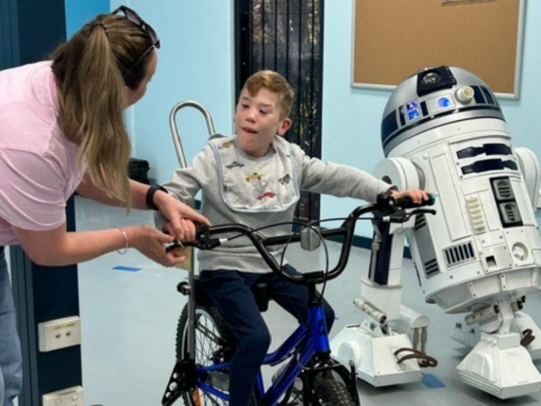 A young boy is sitting on a bike, supported by a woman adjusting the handlebars. The boy is looking at a life-sized R2-D2 robot from Star Wars, which is standing next to him. The scene takes place indoors, and both the boy and the woman are smiling.