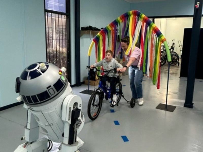 A boy on a tricycle, guided by a woman, rides through a colourful archway made of ribbons. An R2-D2 replica robot is positioned in front of them, creating a playful and engaging atmosphere.