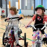 Two young girls are riding adaptive tricycles inside a large indoor sports hall. The girl on the left has glasses and is wearing a striped shirt and floral pants, while the girl on the right is wearing a colourful helmet, a striped shirt, and striped leggings, smiling as they ride side by side.