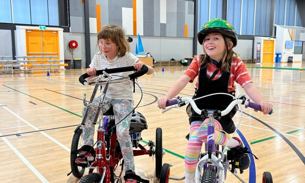 Two young girls are riding adaptive tricycles inside a large indoor sports hall. The girl on the left has glasses and is wearing a striped shirt and floral pants, while the girl on the right is wearing a colourful helmet, a striped shirt, and striped leggings, smiling as they ride side by side.