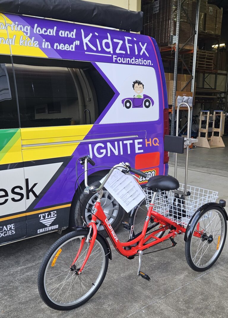 A red adaptive tricycle is parked in front of a colourful van with the branding of KidzFix Foundation and IGNITE HQ. The tricycle has a basket at the back and an information sheet attached to the handlebars. The background shows a warehouse setting with various equipment.