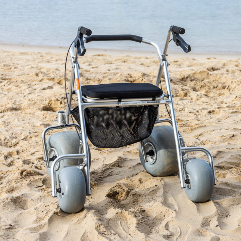 A foldable walker with large, balloon-like wheels designed for beach use is shown on sandy terrain near the water. The walker features a cushioned seat and a black mesh basket for storage.