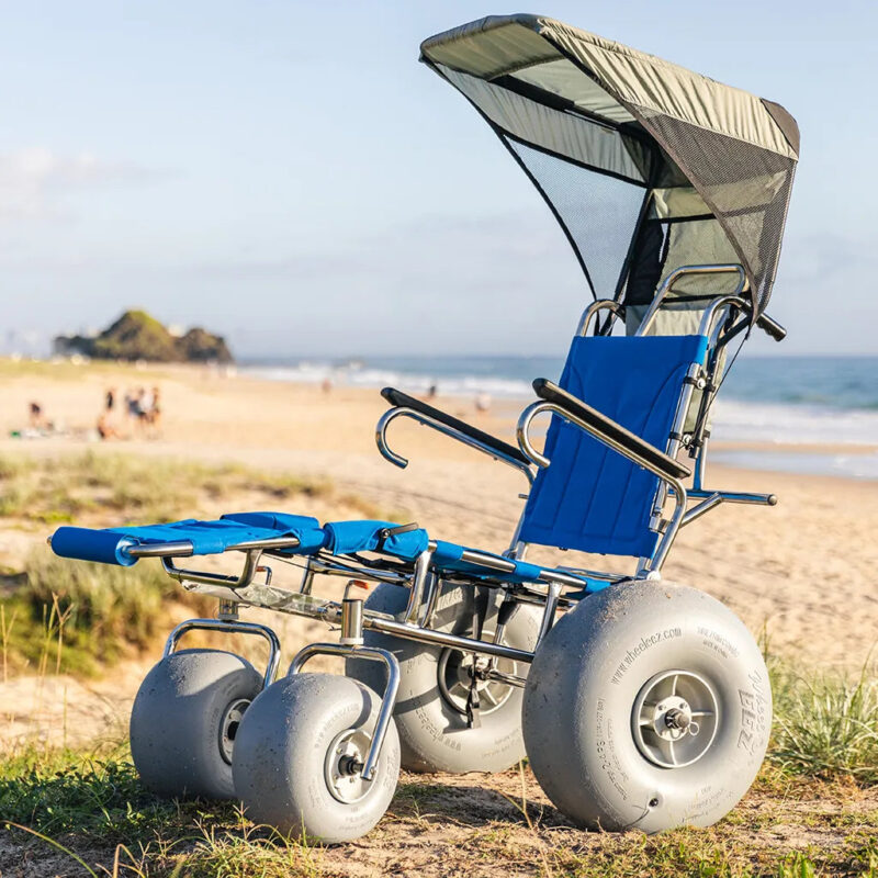 A beach wheelchair with large, balloon-like wheels is positioned on a sandy beach. The wheelchair features a blue reclining seat and an overhead sunshade. The background includes grassy dunes, the ocean, and a distant rocky outcrop, with a few people visible on the beach.