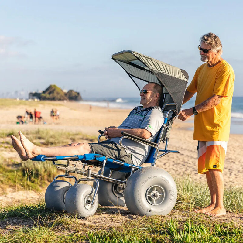 An elderly man relaxes in a beach wheelchair with large balloon wheels and a sunshade, being pushed by another man wearing sunglasses and a yellow shirt. They are on a sandy beach with grass and dunes in the background, enjoying a sunny day with the ocean waves visible in the distance