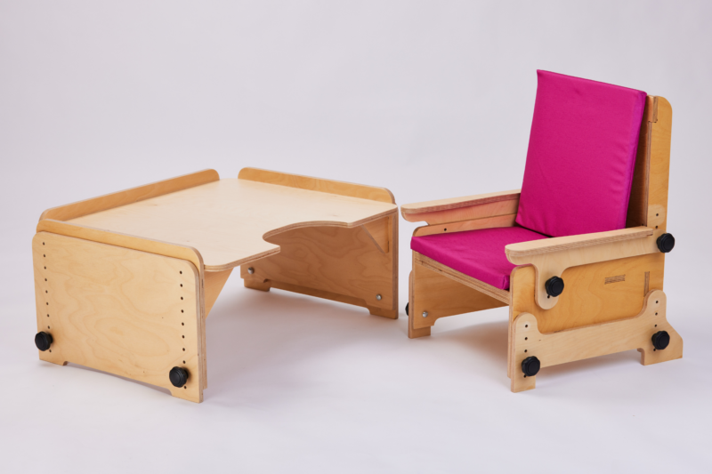 A wooden adjustable desk paired with a matching chair, both featuring a natural finish. The desk has a curved cut-out at the front for ergonomic comfort and black knobs on the sides for height adjustment. The chair, designed to match the desk, has a pink cushioned seat and backrest, providing comfort and support. The simple and functional design emphasizes versatility and ease of use