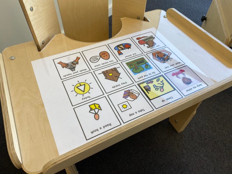 A full view of a wooden desk designed for children with special needs, featuring a communication board with various pictograms under a clear surface. The pictograms depict daily activities such as 'Wash your hands,' 'Eat dinner,' 'Go to the park,' and 'Read a book.' The desk has a curved cut-out for easy accessibility and side rails to prevent items from falling off.