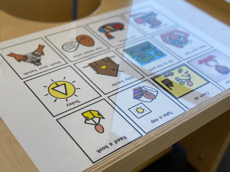 A close-up view of a wooden desk designed for children with special needs, featuring a communication board with various pictograms under a clear surface. The pictograms include images and text for daily activities such as 'Wash your hands,' 'Today,' 'Take a nap,' and 'Read a book