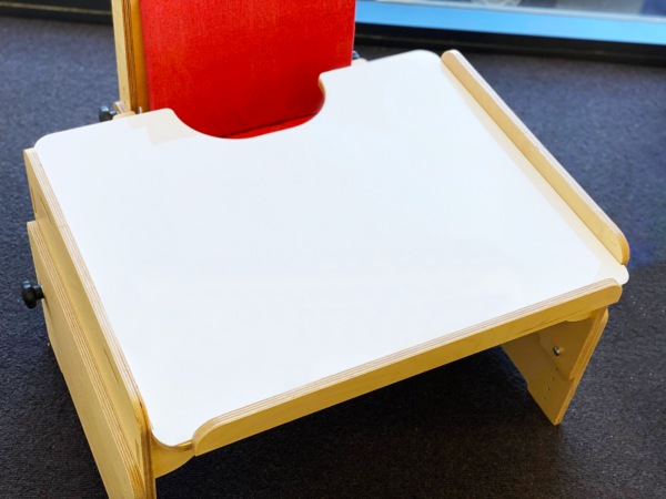 A wooden adjustable chair with a white desk attached, designed for children with special needs. The chair has a red cushioned backrest and side supports to ensure comfort and stability."