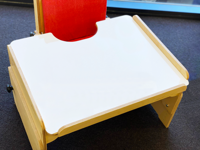 A wooden adjustable chair with a white desk attached, designed for children with special needs. The chair has a red cushioned backrest and side supports to ensure comfort and stability."