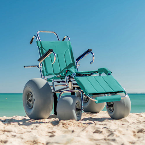 A green beach wheelchair with large balloon wheels sits empty on the sandy shore with a calm, turquoise sea in the background under a clear blue sky."