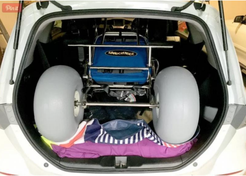 A beach wheelchair with large balloon wheels is neatly packed into the back of a white SUV. The wheelchair, labelled 'Sandcruiser', is folded to fit into the trunk space, with colourful towels or blankets placed underneath