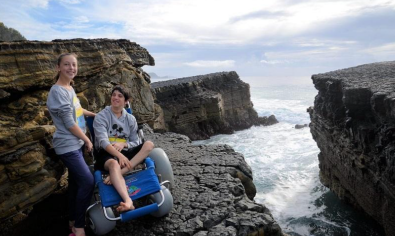 A young man with dark hair, sitting in a beach wheelchair with large balloon wheels, smiles as he looks up at a woman standing beside him. They are on a rocky cliffside overlooking the ocean, with waves crashing against the rocks below. The woman, also smiling, is dressed casually in a grey sweatshirt and blue pants. The scene captures a dramatic coastal landscape under a partly cloudy sky.