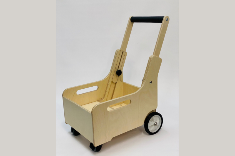 A wooden mobility aid cart with a sturdy rectangular box design, featuring cut-out handles on the sides for easy lifting. The cart has two larger wheels at the rear and two smaller wheels at the front, with a black cushioned handlebar for pushing