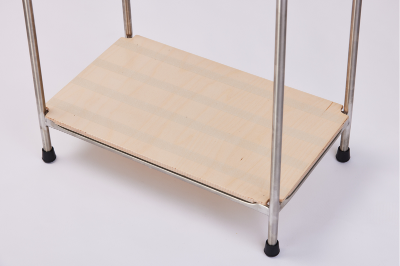 A close-up image of the base of a metal frame stand, featuring a flat wooden platform supported by metal legs. The legs have black rubber caps for stability, and the platform has a striped pattern. The background is plain white