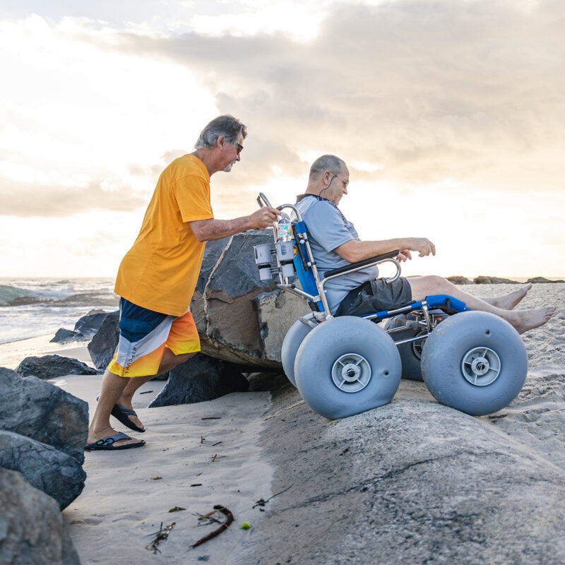 An older man in a grey T-shirt and black shorts is sitting in a beach wheelchair with large balloon wheels, being pushed by another man in an orange T-shirt and multicoloured shorts. They are navigating over sandy terrain near large rocks, with the ocean and a cloudy sky in the background.