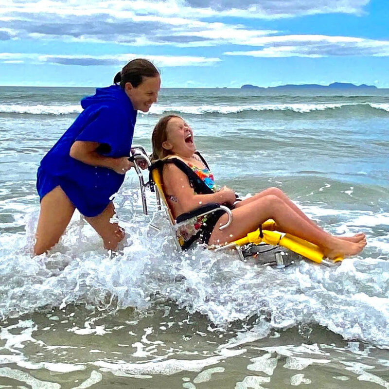 "A young woman pushing a girl in a beach wheelchair with large balloon wheels through the waves at a beach. The girl is laughing joyfully as the water splashes around them. The scene is bright and lively, with a clear sky and distant landforms visible on the horizon.