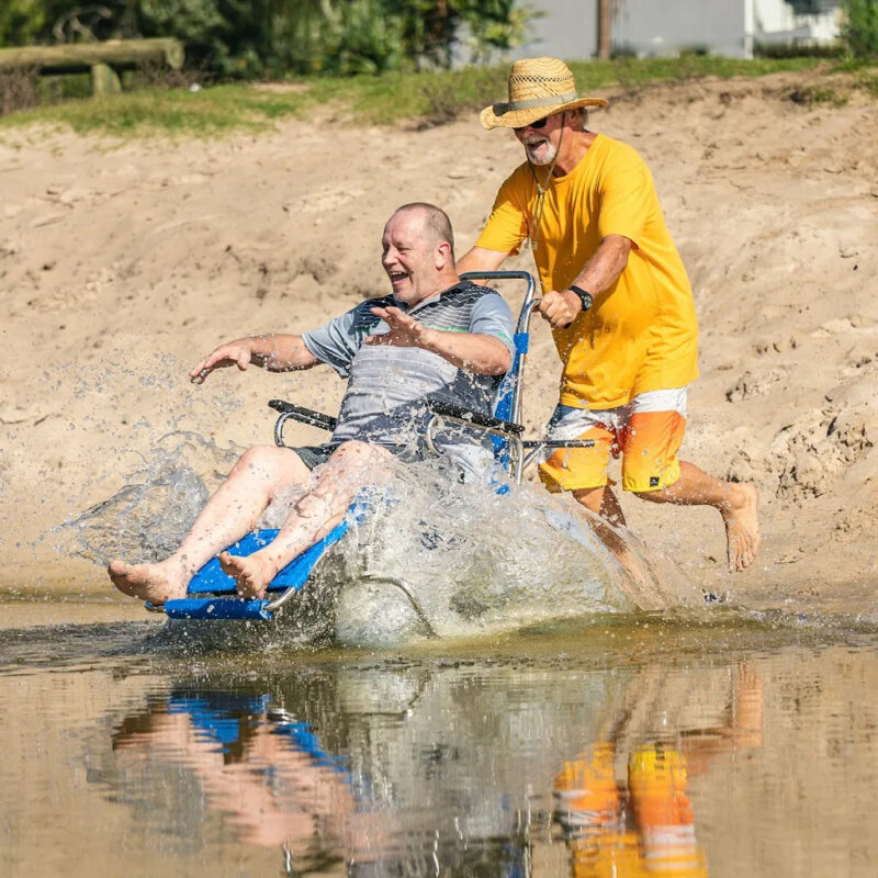 "An elderly man enjoys a splash as he is pushed into the water in a beach wheelchair with large balloon wheels by another man wearing a yellow shirt and a straw hat. They are at a sandy beach area with grass and trees in the background, both smiling and having fun