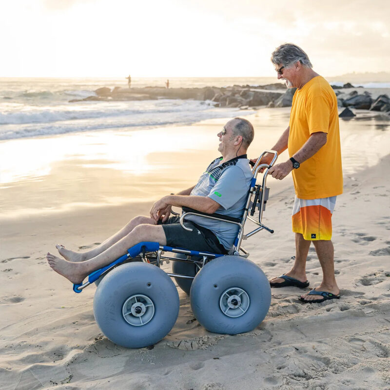 An older man in a grey T-shirt and black shorts is sitting in a beach wheelchair with large balloon wheels. He is being pushed along the beach by another man in an orange T-shirt and multicoloured shorts. The scene is set against a backdrop of the ocean waves and a cloudy sky at sunset