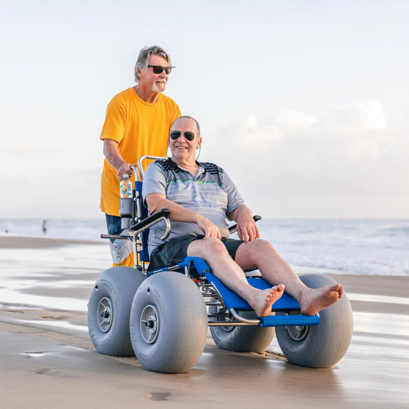 "An older man in a grey polo shirt and black shorts sits comfortably in a beach wheelchair with large balloon wheels, smiling as he enjoys the beach. Another man in an orange T-shirt and sunglasses stands behind him, pushing the wheelchair. The scene captures a serene beach setting with gentle waves and a light, clear sky