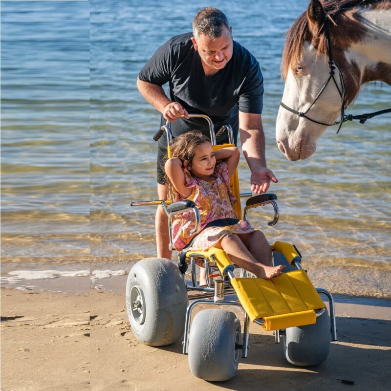 A man in a black T-shirt is pushing a young girl in a yellow beach wheelchair with large balloon wheels on the sand near the water's edge. The girl is wearing a pink dress and is smiling, with her hands behind her head. They are interacting with a large horse standing close to them on the beach.