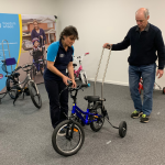 A woman in a "Freedom Solutions" polo shirt is adjusting the handlebars of a small adaptive bike while an older man holds a push handle attached to the bike. They are in a room filled with various adaptive bicycles, with large banners in the background displaying the "Freedom Solutions" and "Freedom Wheels" logos and images of children using adaptive bikes. The setting appears to be a workshop or demonstration area for customising assistive bikes.