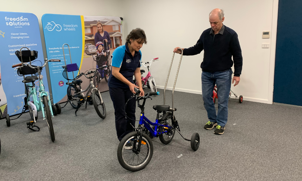 A woman in a "Freedom Solutions" polo shirt is adjusting the handlebars of a small adaptive bike while an older man holds a push handle attached to the bike. They are in a room filled with various adaptive bicycles, with large banners in the background displaying the "Freedom Solutions" and "Freedom Wheels" logos and images of children using adaptive bikes. The setting appears to be a workshop or demonstration area for customising assistive bikes.