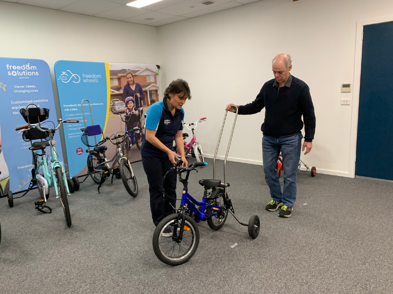 A woman in a "Freedom Solutions" polo shirt is adjusting the handlebars of a small adaptive bike while an older man holds a push handle attached to the bike. They are in a room filled with various adaptive bicycles, with large banners in the background displaying the "Freedom Solutions" and "Freedom Wheels" logos and images of children using adaptive bikes. The setting appears to be a workshop or demonstration area for customising assistive bikes.