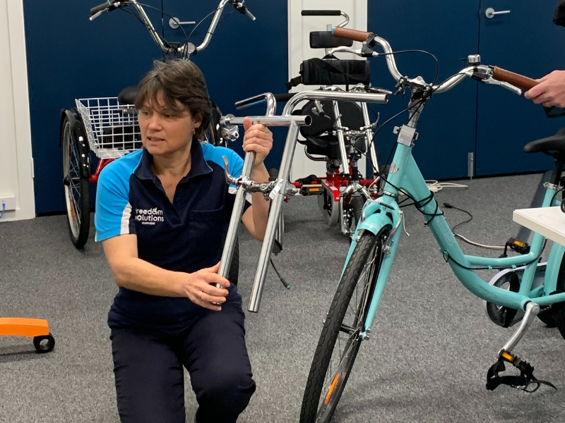 A woman wearing a "Freedom Solutions" polo shirt is kneeling on the floor, holding the handlebars of a bicycle. The setting appears to be a workshop or demonstration area with various adaptive bicycles visible in the background. The woman seems to be in the process of adjusting or assembling the bike. Behind her, there is a red tricycle and another bicycle, indicating a focus on customised or adaptive cycling solutions.