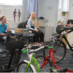 A group of four people is seated in a classroom or workshop setting, attentively listening to an instructor who is explaining modifications on adaptive bicycles. The instructor, wearing a "Freedom Solutions" shirt, is standing beside a row of various bikes, gesturing as they speak. The attendees are seated casually on chairs or stools, with an open laptop in the background displaying a virtual participant. Posters of human anatomy are visible on the wall, indicating a clinical or educational environment