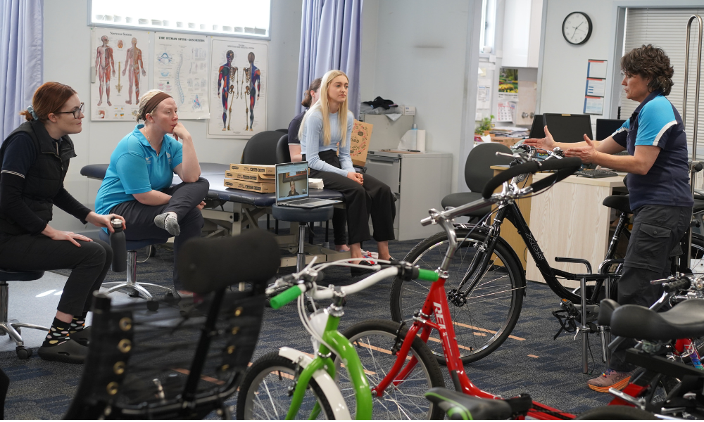 A group of four people is seated in a classroom or workshop setting, attentively listening to an instructor who is explaining modifications on adaptive bicycles. The instructor, wearing a "Freedom Solutions" shirt, is standing beside a row of various bikes, gesturing as they speak. The attendees are seated casually on chairs or stools, with an open laptop in the background displaying a virtual participant. Posters of human anatomy are visible on the wall, indicating a clinical or educational environment