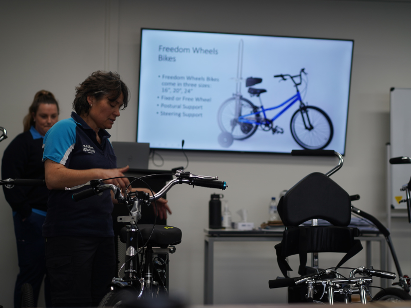 A staff member from Freedom Solutions Australia is demonstrating an adaptive bicycle in an indoor setting. The person is wearing a navy blue and teal uniform, and they are adjusting the bike’s handlebars. Behind them, a presentation is displayed on a screen, showing a Freedom Wheels bike with details about various bike sizes, wheel options, and postural support. Another staff member stands in the background, assisting with the session. The environment appears to be a training or informational workshop focused on adaptive cycling for people with disabilities.