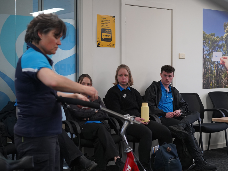 A staff member from Freedom Solutions Australia is demonstrating the handlebars of a red adaptive bicycle during an indoor session. Several participants, wearing blue uniforms, are seated in the background, attentively watching the demonstration. The environment suggests a classroom or training setting. A yellow mental health awareness poster is visible on the wall behind the participants, promoting the message "R U OK?" to encourage mental wellness. The participants appear engaged as they observe the session focused on adaptive cycling solutions for people with disabilities.