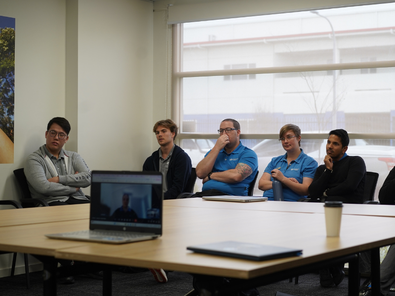 A group of five individuals sit around a conference table, attentively listening to a presentation. Two of them wear blue Freedom Solutions Australia polo shirts, while the others are casually dressed. A laptop and coffee cup are placed on the table in front of them. The group appears focused, with some participants resting their hands on their chins in thought. Behind them, large windows offer a view of an industrial building outside. The setting suggests a training or meeting session.