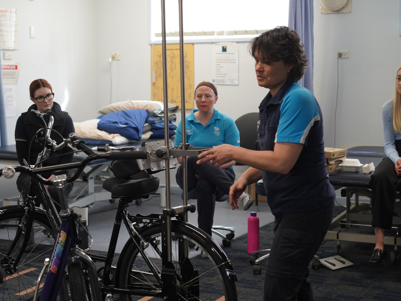 A woman in a Freedom Solutions Australia polo shirt demonstrates the adjustments on a modified bicycle while two attendees, one wearing a blue polo and the other in a dark outfit, observe attentively. The modified bicycle includes additional support structures and adaptive components. The room appears to be a clinical or therapy setting with physical therapy tables and pillows in the background. The setup suggests a hands-on demonstration of assistive technology for individuals with mobility challenges.