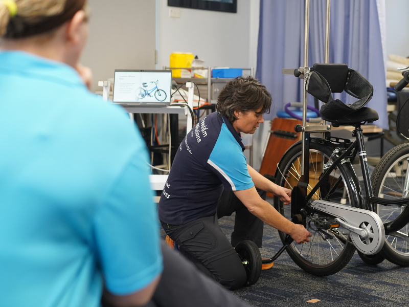 A woman in a Freedom Solutions Australia polo shirt kneels on the floor, adjusting the support structures on a modified black bicycle. The bicycle features adaptive components, including a backrest and stabilisers, indicating its use for individuals with specific mobility or posture needs. The environment appears to be a clinical or therapy room, with medical equipment and curtains visible in the background. The woman is focused on the adjustment, demonstrating the customisation process of assistive technology.